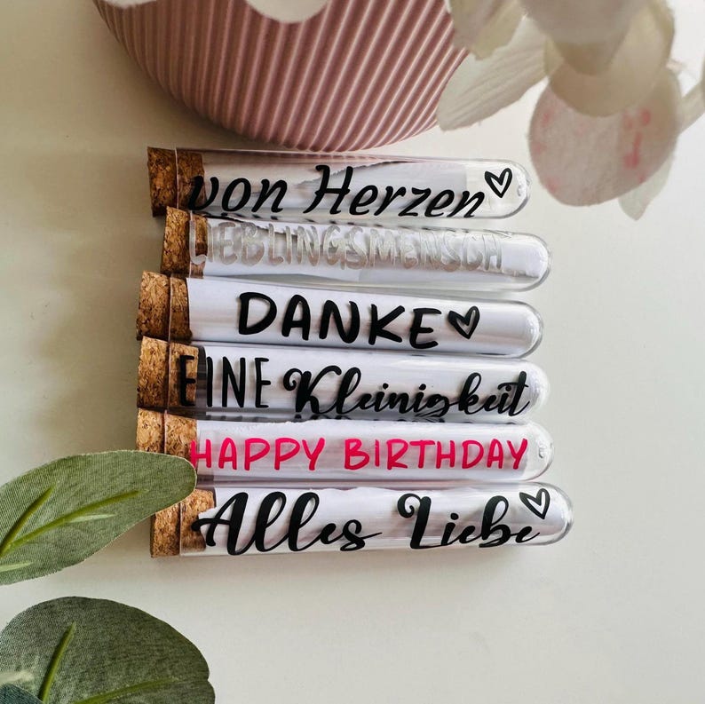 May include: A stack of glass tubes with cork stoppers, each containing a rolled-up message. The messages are written in black and pink script, including "von Herzen", "LIEBLINGSMENSCH", "DANKE", "EINE Kleinigkeit", "HAPPY BIRTHDAY", and "Alles Liebe".