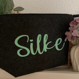 Könnte beinhalten: Eine graue Filz-Kosmetiktasche mit dem Namen "Silke" in grüner Schrift.