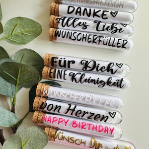 Könnte beinhalten: Eine Sammlung von Glasröhrchen mit Korken, die jeweils eine gerollte Nachricht enthalten. Die Röhrchen sind mit verschiedenen deutschen Sätzen beschriftet, darunter "LIEBLINGSMENSCH" und "HAPPY BIRTHDAY", in schwarzer, silberner und goldener Schrift.