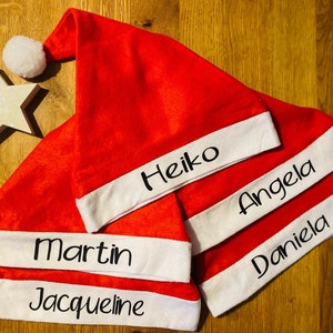 Könnte beinhalten: Rote Weihnachtsmützen mit weißem Rand, personalisiert mit Namen in schwarzer Schrift. Die Mützen sind auf einer Holzoberfläche angeordnet, mit einer sternförmigen weißen Dekoration und einem weißen Bommel.