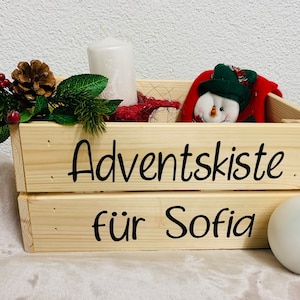 May include: A wooden crate with the words "Adventskiste für Sofia" written on it. The crate is filled with Christmas decorations, including a snowman, a pine cone, and a candle.