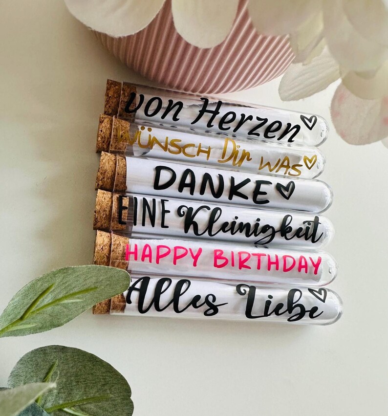 May include: A collection of glass tubes with cork stoppers, each labeled with a different message in black or gold script. The messages include "Von Herzen", "Wunsch Dir Was", "Danke", "Eine Kleinigkeit", "Happy Birthday", and "Alles Liebe".