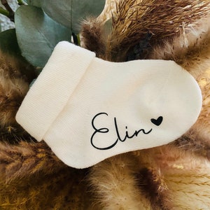 May include: White baby sock with the name "Elin" written in black cursive with a small black heart next to it.