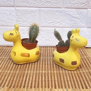 May include: Two yellow giraffe-shaped ceramic planters, each holding a small cactus in a brown pot. The planters have brown spots and are set on a bamboo mat against a white brick wall.