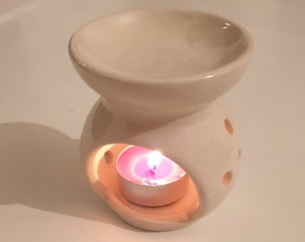 May include: White ceramic oil diffuser with a round, open top and a small, round opening on the side. A small, white tea light candle is burning inside the opening, casting a warm glow.