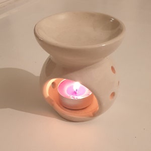 Op de afbeelding: Witte keramische olieverdamper met een ronde, open bovenkant en een kleine, ronde opening aan de zijkant. Een kleine, witte theelichtje brandt in de opening en geeft een warm licht.