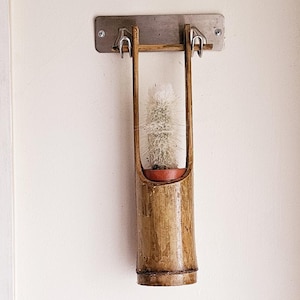 Op de afbeelding: Een hangende plantenhanger gemaakt van bamboe met een kleine cactusplant erin. De plantenhanger is bevestigd aan een metalen beugel die aan een witte muur is gemonteerd.