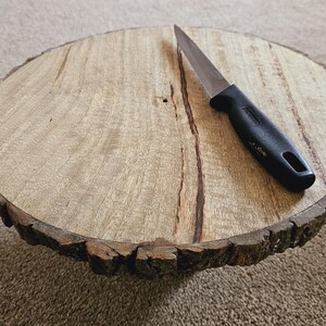 Puede incluir: Una bandeja redonda de madera para servir con un borde de corteza natural. Un cuchillo con mango negro se coloca en la bandeja.