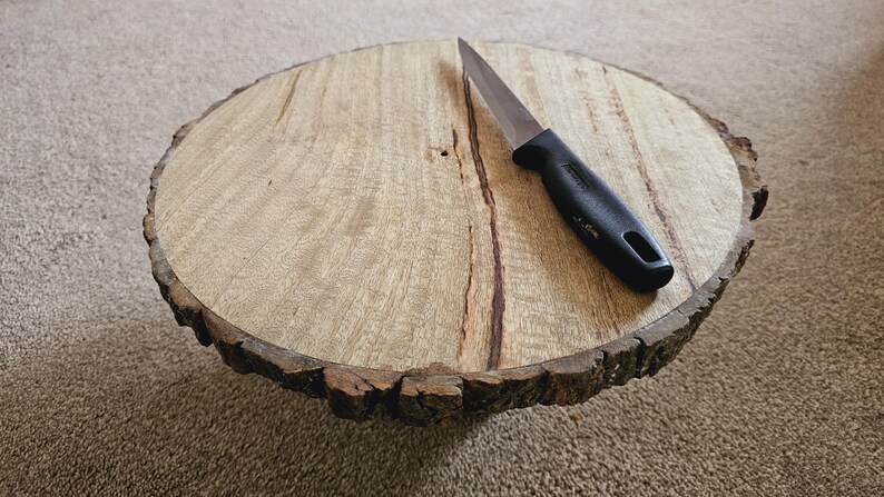 Puede incluir: Una bandeja redonda de madera para servir con un borde de corteza natural. Un cuchillo con mango negro se coloca en la bandeja.