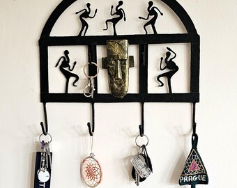 May include: Black metal wall hanging with six hooks, featuring tribal dancer silhouettes and a central mask design. The text reads "Handcrafted Tribal Relics" and "Bastar Tribal WALL HANGING".