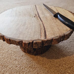 Puede incluir: Un soporte para tartas de madera con un borde de corteza natural y un cuchillo negro en la parte superior.