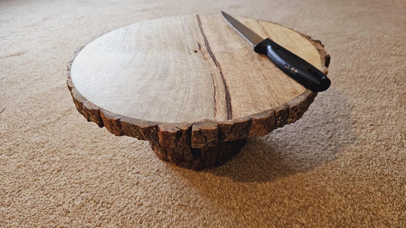 Puede incluir: Un soporte para tartas de madera con un borde de corteza natural y un cuchillo negro en la parte superior.