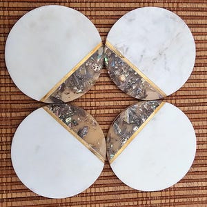 May include: Four round white marble coasters with a gold trim and iridescent shell inlay. The coasters are arranged in a square pattern on a brown woven surface.