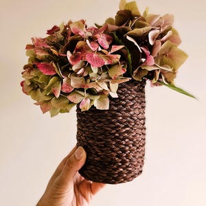 Può includere: Un vaso in vimini intrecciato marrone con un bouquet di ortensie secche in tonalità rosa e verdi.