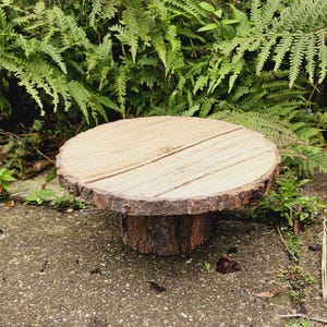 Puede incluir: Un soporte para tartas de madera r&uacute;stico con un borde de corteza natural y una parte superior lisa de color marr&oacute;n claro. El soporte est&aacute; sobre una superficie de hormig&oacute;n gris.