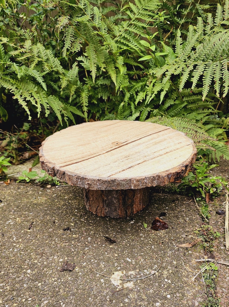 Puede incluir: Un soporte para tartas de madera r&uacute;stico con un borde de corteza natural y una parte superior lisa de color marr&oacute;n claro. El soporte est&aacute; sobre una superficie de hormig&oacute;n gris.