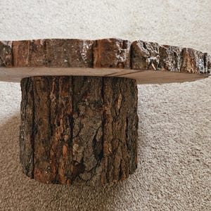 Puede incluir: Un soporte para tartas de madera con un acabado natural de corteza. El soporte est&aacute; hecho de una rebanada de tronco de &aacute;rbol y tiene una parte superior redonda.