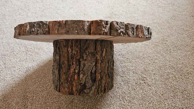 Puede incluir: Un soporte para tartas de madera con un acabado natural de corteza. El soporte est&aacute; hecho de una rebanada de tronco de &aacute;rbol y tiene una parte superior redonda.
