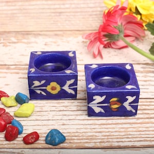 May include: Two blue ceramic candle holders with white and yellow floral designs. The holders are square shaped and have a small round opening in the centre for a candle.