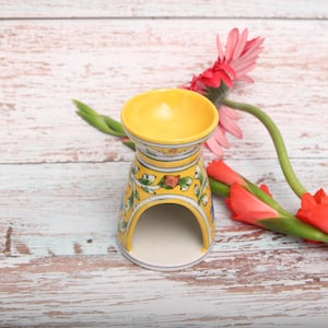 Può includere: Un bruciatore di oli essenziali in ceramica gialla con base svasata e parte superiore a forma di ciotola. L'esterno presenta un motivo floreale dipinto a mano con foglie verdi e fiori arancioni su fondo giallo, con sottili linee blu.