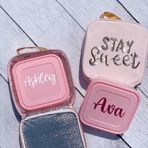 Op de afbeelding: Twee roze lunchtassen met geïsoleerde binnenkant. Eén tas heeft de tekst "Stay Sweet" in zilveren glitters. De andere tas heeft de naam "Ashley" in witte tekst. De derde tas heeft de naam "Ava" in roze tekst.