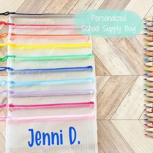 May include: A set of eight clear zippered mesh bags with rainbow colored zippers. The bags are stacked on top of each other and have a personalized name "Jenni D." on the bottom bag. The bags are on a wood background.  The text "Personalized School Supply Bag" is written in blue on a light blue background.