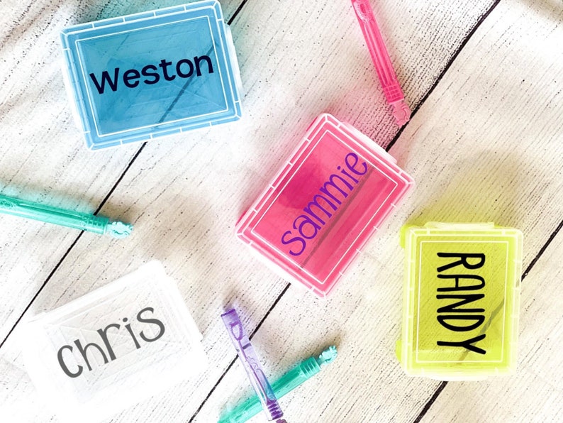 Op de afbeelding: Vier doorzichtige plastic opbergdozen met kleurrijke deksels. De dozen zijn gelabeld met de namen "Weston", "Sammie", "Randy" en "Chris".
