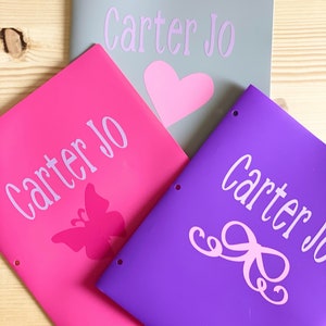 May include: Three personalized binders with the name "Carter Jo" printed on each. The binders are gray, pink, and purple. The pink binder has a butterfly design, the purple binder has a bow design, and the gray binder has a heart design.