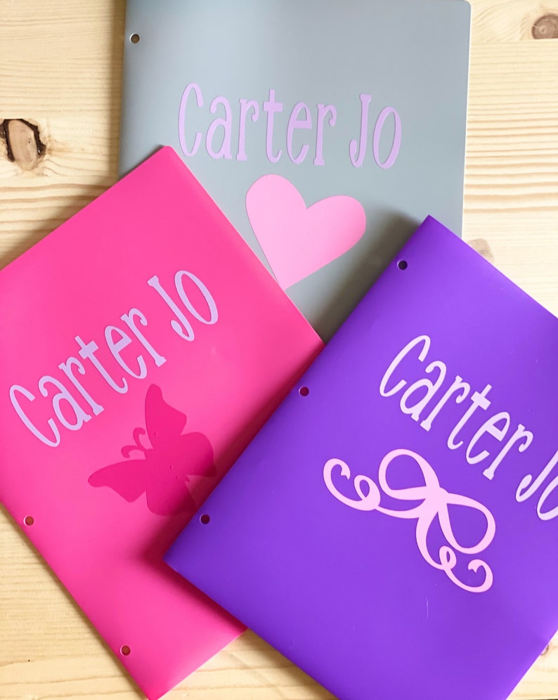 May include: Three personalized binders with the name "Carter Jo" printed on each. The binders are gray, pink, and purple. The pink binder has a butterfly design, the purple binder has a bow design, and the gray binder has a heart design.