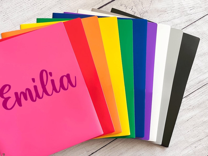 May include: A stack of colorful plastic folders with the name "Emilia" written on the front of the pink folder.
