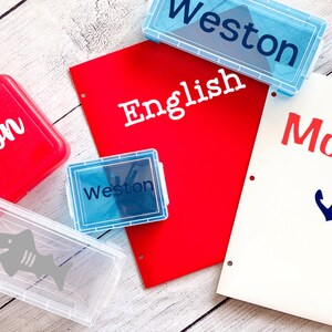 May include: Four clear plastic pencil boxes with different designs. One box is red with the name "Wilson" in white lettering. Another box is blue with the name "Weston" in white lettering. A third box is clear with a shark design. The fourth box is blue with the name "Weston" in white lettering. A red folder with the word "English" in white lettering is visible in the background.