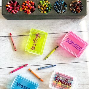 Op de afbeelding: Vier doorzichtige plastic containers met deksels om kleurpotloden in op te bergen. Elke container heeft een deksel in een andere kleur en een naam erop geschreven:  "Weston" in blauw, "Emilia" in wit, "Sammie" in roze en "Brileigh" in geel. De containers staan op een wit houten oppervlak.