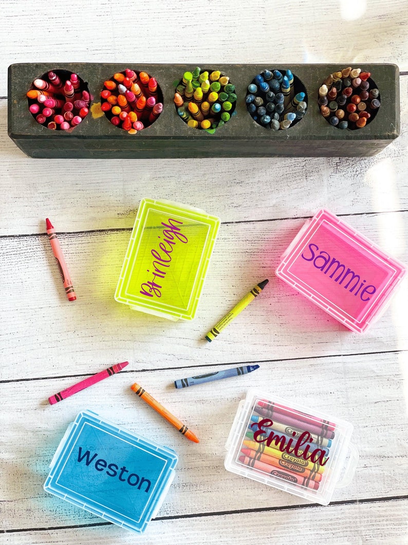 Op de afbeelding: Vier doorzichtige plastic containers met deksels om kleurpotloden in op te bergen. Elke container heeft een deksel in een andere kleur en een naam erop geschreven:  "Weston" in blauw, "Emilia" in wit, "Sammie" in roze en "Brileigh" in geel. De containers staan op een wit houten oppervlak.