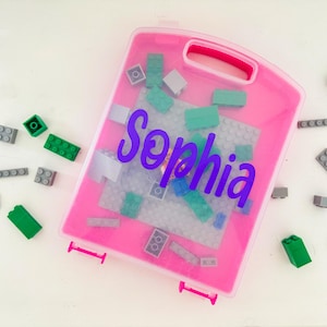 May include: A pink plastic storage case with a handle, containing various colored toy blocks. The name "Sophia" is written in purple script on the case. Several blocks are scattered around the case on a white surface.