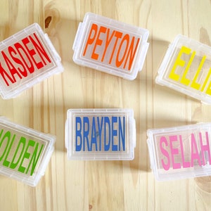 Op de afbeelding: Zes doorzichtige plastic containers met kleurrijke labels. De labels hebben de namen Kasden, Peyton, Ellie, Brayden, Selah en Holden.