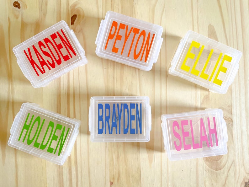 Op de afbeelding: Zes doorzichtige plastic containers met kleurrijke labels. De labels hebben de namen Kasden, Peyton, Ellie, Brayden, Selah en Holden.