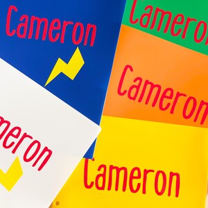 May include: Five colorful folders with the name "Cameron" written in red on each folder. The folders are blue, green, orange, yellow, and white. The blue, white, and yellow folders have a yellow lightning bolt graphic.