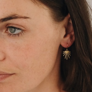 May include: A close-up of a person wearing gold sunburst earrings. The earrings are small and delicate, with a simple design.