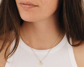 Könnte beinhalten: Eine Frau trägt ein weißes Tanktop mit einer goldenen Kettenkette und einem kleinen goldenen Kreisanhänger.