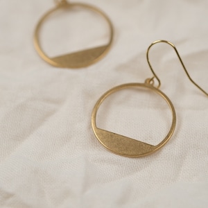 Könnte beinhalten: Ein Paar goldfarbene Ohrringe mit einem schlichten, modernen Design. Jeder Ohrring hat eine kreisförmige Form mit einem horizontalen Balken in der Mitte.