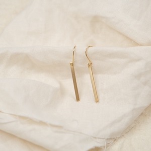 Könnte beinhalten: Ein Paar goldene Bar-Ohrringe mit einem schlichten, minimalistischen Design. Die Ohrringe bestehen aus einem dünnen, rechteckigen Goldstab, der an einem kleinen, filigranen Haken hängt.