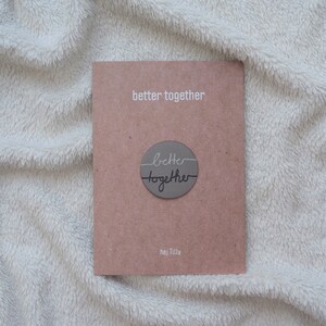 Può includere: Un biglietto marrone con il testo "better together" in lettere bianche. Un cerchio grigio con il testo "better together" in lettere nere è centrato sul biglietto. Il testo "hej Tilly" è stampato in bianco nella parte inferiore del biglietto.