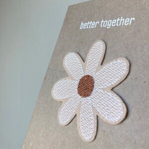 Könnte beinhalten: Eine braune Karte mit dem Text "better together" in weiß gedruckt. Eine weiße gestickte Gänseblümchen mit brauner Mitte ist an der Karte befestigt.