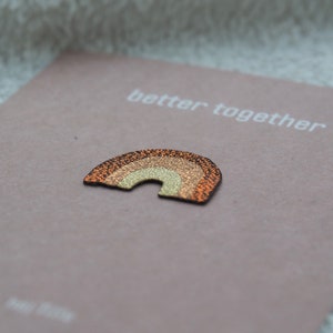 Könnte beinhalten: Ein kleiner, glitzernder Regenbogen-förmiger Schmuck in Orange-, Gold- und Brauntönen, der auf einer rosa Karte liegt. Auf der Karte steht in Weiß "better together".