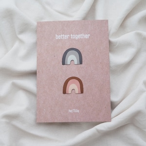 Könnte beinhalten: Eine rosa Karte mit dem Text "better together" darauf gedruckt. Zwei regenbogenförmige Papierschnitte sind an der Karte befestigt. Der obere Regenbogen ist grau und weiß, der untere Regenbogen ist braun, rosa und weiß. Der Text "hej Tilly" ist unten auf der Karte gedruckt.