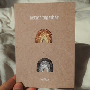 Könnte beinhalten: Eine braune Karte mit dem Text "better together" und zwei glitzernden Regenbogen-Ansteckern. Der obere Regenbogen hat Gold- und Bronzetöne, während der untere silber- und graufarben ist. Die Karte trägt auch den Text "hej Tilly".