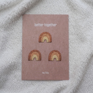 Könnte beinhalten: Drei braune und goldene Glitzer-Regenbogen-Patches auf einer hellbraunen Karte mit dem Text "better together" und "hej Tilly".