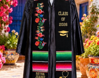May include: A black graduation gown worn with a black graduation stole featuring colorful serape-style stripes at the ends, floral embroidery, and gold text reading "CLASS OF 2026", "REYNA JUAREZ DOROTEO", a graduation cap icon, and a stethoscope icon.
