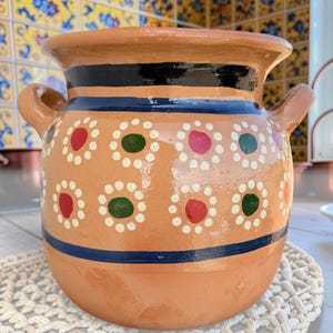 May include: A terracotta pot with two side handles, featuring a wide rim and a smooth finish. The exterior is decorated with horizontal black and blue bands and a repeating pattern of white-dotted circles containing red or green centers.