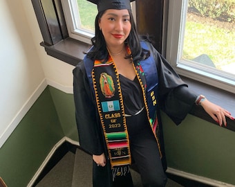 May include: A person wearing a black graduation gown and cap, a blue and gold sash, and a colorful graduation stole with the text "Class of 2023" and a flag of Guatemala. The person is standing on a staircase.